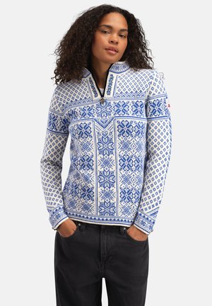 Kvinne med krøllete hår iført en hvit og blåmønstret zip-up genser og svarte bukser, med hendene i lommene, foran en enkel bakgrunn.