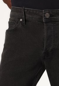 Jeans de mezclilla negra con una textura suave, cierre de botón, bolsillos frontales y presillas para cinturón. Presenta un ajuste clásico y sin patrones visibles.