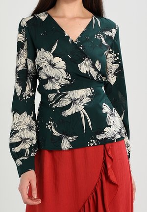 Femme portant un chemisier portefeuille vert foncé à motifs floraux blancs et une jupe rouge avec un bord froncé.