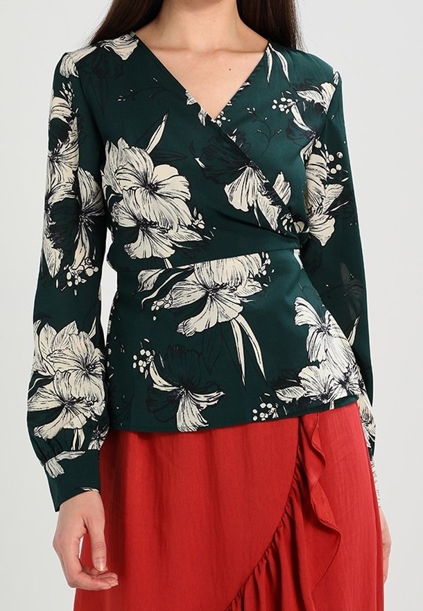Femme portant un chemisier portefeuille vert foncé à motifs floraux blancs et une jupe rouge avec un bord froncé.