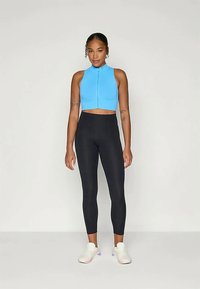 Kvinne som står iført en ermeløs, blå cropped zip-up topp, høyt liv svarte leggings, og hvite joggesko på en enkel bakgrunn.