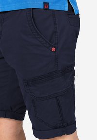 Navy cargo shorts made from textured fabric, featuring two front pockets, one side pocket with flap, and a red accent button.