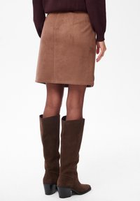 Vrouw in een bruine suède rok, transparante panty’s en donkerbruine suède laarzen tot de knie, staande tegen een effen witte achtergrond.
