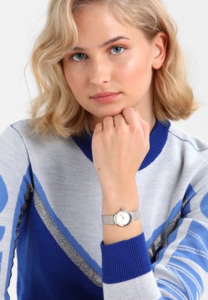 Giovane donna con capelli biondi che indossa un maglione blu e grigio, poggia il mento sulla mano, mostrando un orologio da polso d'argento.