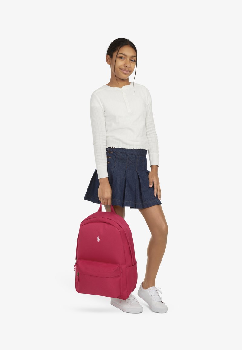 Sac à dos rouge avec un extérieur en tissu lisse, un compartiment principal unique, une poche frontale et un logo blanc. Le modèle porte un haut blanc et une jupe en jean.