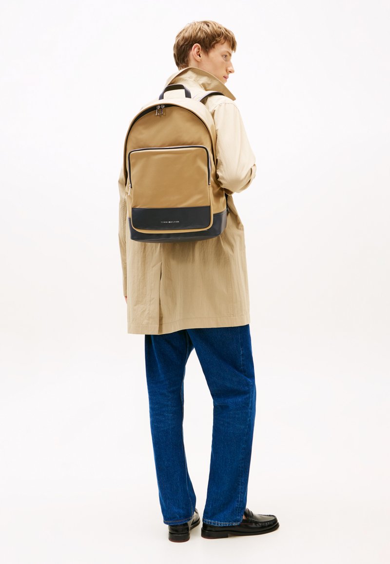 Zaino in nylon marrone con texture liscia, dettagli neri sul fondo e una tasca frontale con zip. Indossato sopra un cappotto beige e jeans blu.