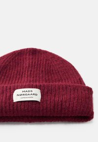 Gorro de malha em lã escura burdeos com textura canelada. Apresenta um rótulo branco com "MADS NØRGAARD COPENHAGEN" em texto preto.