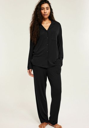 Vrouw met lang krullend haar draagt een zwarte, knoopsluitende top met lange mouwen en bijpassende losse broek, staat op blote voeten tegen een effen achtergrond.