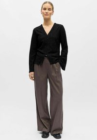 Vrouw staand, gekleed in een zwarte gestructureerde blouse met knopen, losse grijze broek, zwarte schoenen, met haar naar achteren gebonden, neutrale gezichtsuitdrukking.