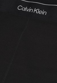 Černý pás značky Calvin Klein s bílým logem, hladkou texturou a minimalistickým designem, s centrálním švem podél těla.