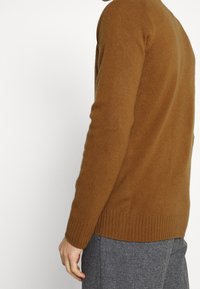 Bruine gebreide trui met geribbelde manchetten en zoom, met een zachte textuur. Gestyled met grijze geruite broek.