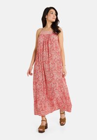 Robe maxi à motifs rouges avec des fines bretelles, tissu froncé au niveau de la poitrine et une silhouette fluide. Associée à des sandales à plateforme marron.