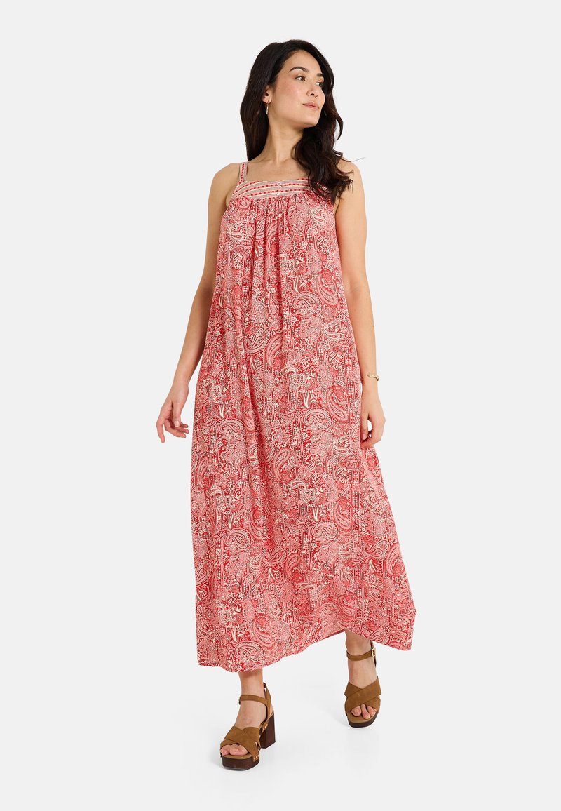 Robe maxi à motifs rouges avec des fines bretelles, tissu froncé au niveau de la poitrine et une silhouette fluide. Associée à des sandales à plateforme marron.