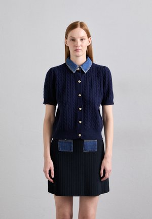 Marinblå kabelstickad cardigan med korta ärmar, denimkrage och guldknappaccent. Matchad med en ribbad svart kjol med denimfickor.