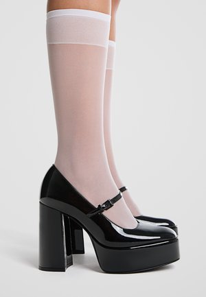 Chaussures à talons hauts en cuir verni noir avec une plateforme épaisse et une bride à la cheville, assorties à des bas blancs transparents montant jusqu'aux genoux.