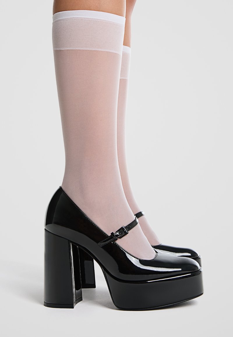 Chaussures à talons hauts en cuir verni noir avec une plateforme épaisse et une bride à la cheville, assorties à des bas blancs transparents montant jusqu'aux genoux.