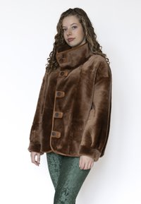 Bruine nepbonten jas met hoge kraag, voorzien van vijf knopen en bruine lederen accenten, gecombineerd met groene getextureerde legging.