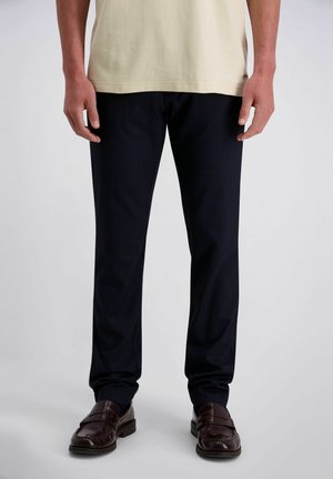 Mann trägt dunkelblaue Hose, beige Hemd und braune Lederloafer, steht vor einem schlichten hellen Hintergrund.