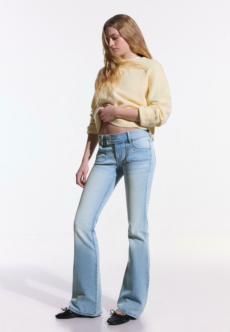 Jeune femme aux longs cheveux blonds, vêtue d'un pull jaune pâle et d'un jean flare bleu clair, debout devant un fond blanc.
