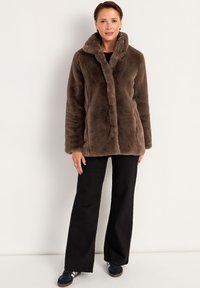 Brun faux fur-jakke med en blød tekstur, notch-krave og lukning foran. Kombineret med sorte bukser med brede ben og sorte sneakers.