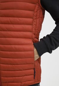Crveni puffer prsluk sa kviltiranim dizajnom i bočnim džepovima. Crni pleteni manžete su vidljive. Glatka tekstura s sjajnom završnom obradom.