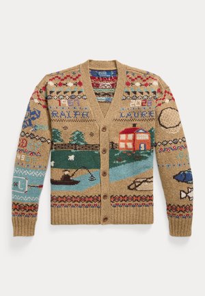 Jeune homme portant un cardigan boutonné à motifs, une chemise à carreaux, une cravate bleu marine, un pantalon beige, des chaussures marron et une casquette de baseball bleu marine, debout avec une main dans la poche.
