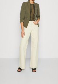 Zielonkawozielona dopasowana kurtka z dwoma przednimi zamkami, noszona na brązowym i czarnym topie w stylu tie-dye, połączona z jasnobeżowymi spodniami w szerokim kroju i czarnymi szpilkami.