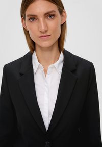 Schwarzer taillierter Blazer mit Revers, einreihigem Knopfverschluss und strukturiertem Stoff, getragen über einem weißen Hemd mit Kragen.