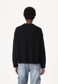 Črn pleten pulover ohlapnega kroja z rebrastim robom in krožnim izrezom, v kombinaciji s svetlo modrimi cargo hlačami. Tekstura se zdi mehak.