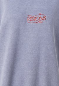 Ljusblå sweatshirt i mjukt material. Har röd broderad text "NYC VISIONS Est. 1995." Enkel design utan synliga mönster.