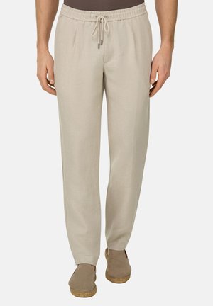 Beige Leinenhose mit Kordelzug, getragen mit braunen Espadrilles zum Hineinschlüpfen, an einer Person stehend vor einem schlichten Hintergrund.