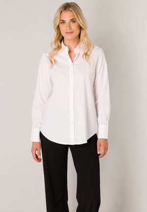 Blonde vrouw die een wit blouse met knopen en een zwarte broek draagt, staand tegen een effen lichtgrijze achtergrond.