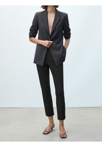 Kvinna som bär en mörkgrå blazer med uppvikt ärmar, svarta slim-fit byxor och svarta öppna klackar, stående mot en enkel bakgrund.