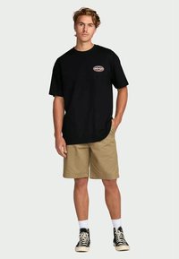 Zwarte katoenen t-shirt met korte mouwen, een afgeronde zoom en een geborduurd logo op de borst, gecombineerd met lichtbruine shorts en zwarte sneakers.