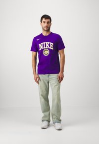 T-shirt en coton violet avec un logo "NIKE" jaune bien visible et un graphique circulaire, associé à un jean décontracté à lavages clairs et des baskets blanches.