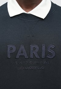 Sudadera azul marino oscuro con un cuello blanco. Presenta texto bordado: "PARIS", dirección, y "KARL LAGERFELD" en colores contrastantes. Tejido suave.