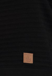 Suéter de punto negro con un patrón de rayas texturizadas y una etiqueta de cuero marrón con el grabado "KEY LARGE" en la parte inferior izquierda.