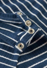 Granatowo-niebieska, w paski tkanina z białymi liniami, z teksturowanym prążkowaniem i metalowymi zatrzaskami wzdłuż dekoltu. Zbliżenie na detal odzieży.