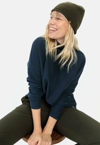 Donkerblauwe geribde trui met een hoge hals, gecombineerd met een olijfgroene aangesloten broek. Het model draagt een bijpassende olijfgroene muts.