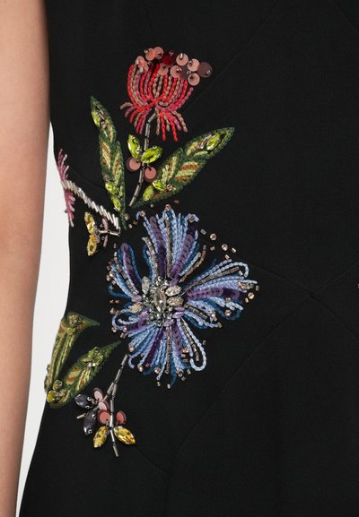 Juodas drabužis su sudėtingu gėlių siuvinėjimu, puoštas spalvotais karoliukais ir blizgučiais. Akcentai – violetinės, raudonos ir žalios spalvos detalės.