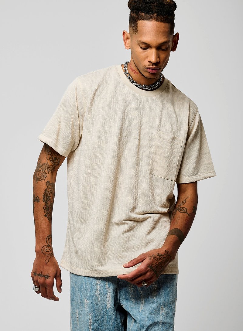 T-shirt beige à manches courtes avec un tissu texturé, col rond et une poche sur la poitrine gauche. Associe à un jean en denim clair usé.