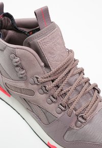 Reebok High-Top-Sneaker in hellem Mauve mit Mesh- und Wildledermaterialien, ausgestattet mit strukturierten Schnürsenkeln, einer Gummisohle und einem roten Akzent an der Ferse.