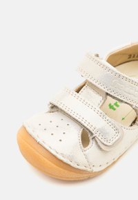 Sølvmetallisk sandal med to justerbare Velcro-stropper, perforert tåområde, og en tan gummisåle, med eksponerte sømdetaljer.