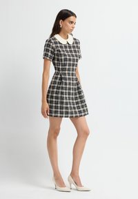 Donna che indossa un vestito a scacchi bianco e nero a maniche corte con un colletto Peter Pan bianco e tacchi alti bianchi, in piedi di lato.