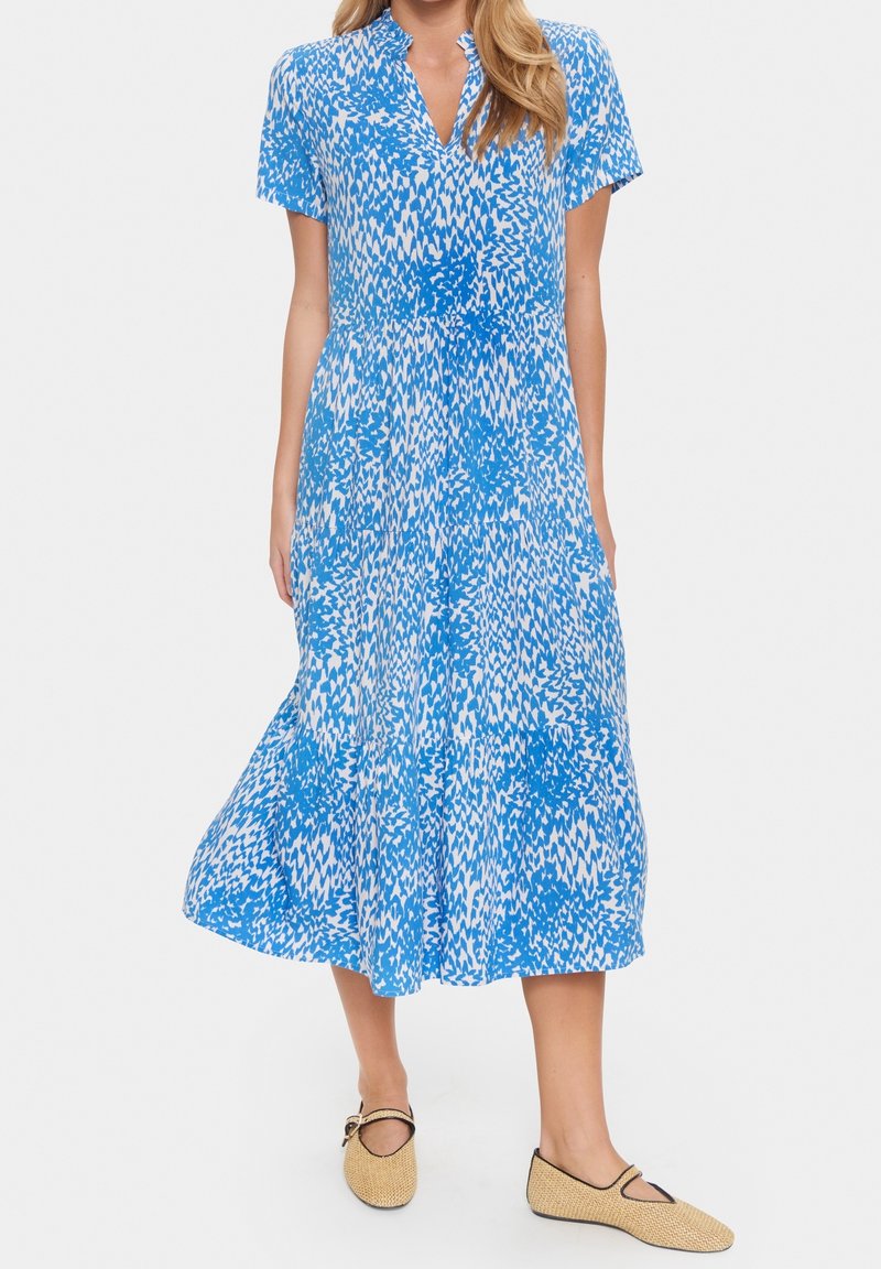 Femme portant une robe midi à manches courtes bleue et blanche à motifs avec un col et des chaussures plates beiges sur un fond uni.