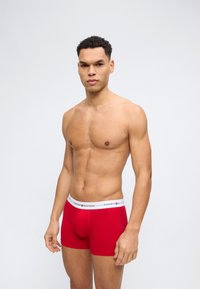 Junger Mann mit kurzen lockigen Haaren steht oberkörperfrei und trägt rote Tommy Hilfiger Boxershorts vor schlichtem hellem Hintergrund.