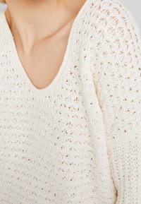 Teksturowany biały sweter dzianinowy z szerokim dekoltem w kształcie litery V i otwartym wzorem dzianiny, podkreślający miękki materiał i luźny krój.