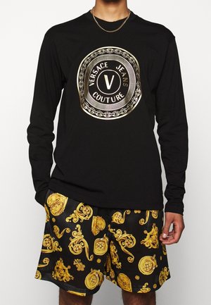 Person trägt ein schwarzes Langarmshirt mit goldenem Versace Jeans Couture Logo und schwarze Shorts mit verzierten goldenen Barockmotiven.