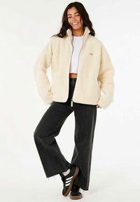 Crèmekleurig kunstbontjack met een ritssluiting aan de voorkant, opstaande kraag en een lichte glans. Gecombineerd met hooggetailleerde zwarte wijde broekspijpen en sneakers.