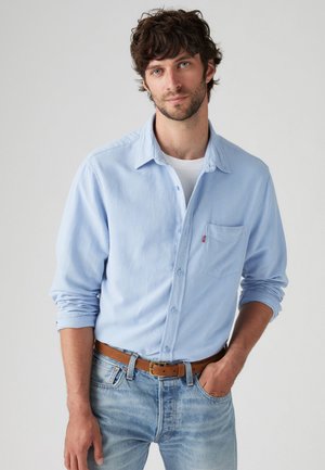 Hellblaues Hemd mit Knopfleiste und Brusttasche, kombiniert mit ausgewaschenen blauen Jeans und einem braunen Gürtel. Das Hemd hat aufgerollte Ärmel.